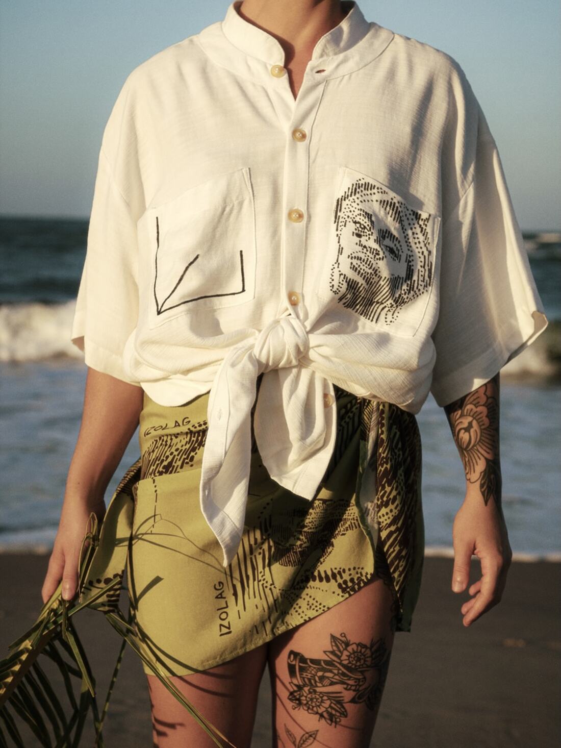 Modelo na praia com camisa off white e saia verde amarrada